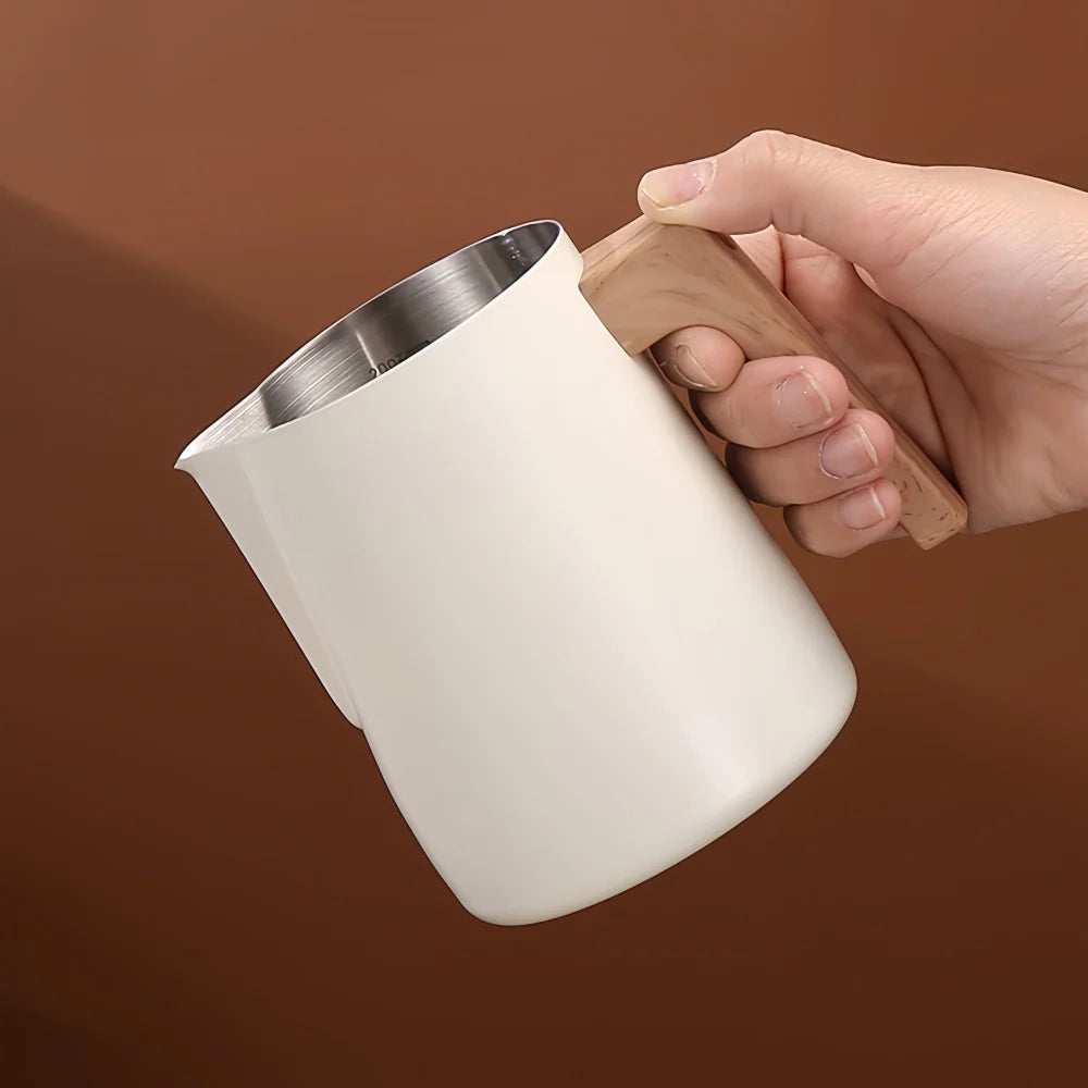 A hand holding a matte off-white milk pitcher with a stainless steel interior and a light brown wooden handle, set against a solid warm brown background.