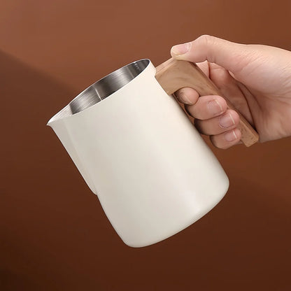 A hand holding a matte off-white milk pitcher with a stainless steel interior and a light brown wooden handle, set against a solid warm brown background.