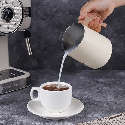 A hand holds a cream-colored stainless steel milk pitcher pouring milk into a white coffee cup on a saucer, with a coffee machine visible in the background.