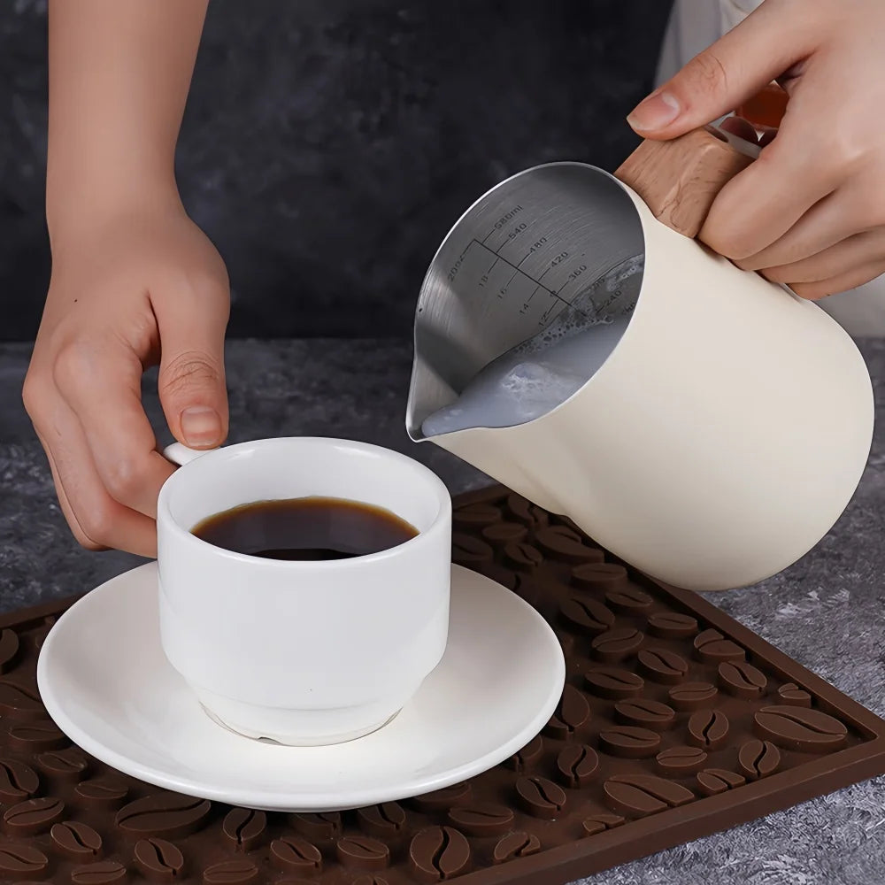 A person pouring milk from a cream-colored stainless steel milk pitcher into a white cup of black coffee on a saucer, set on a brown silicone coffee mat with a dark gray background.