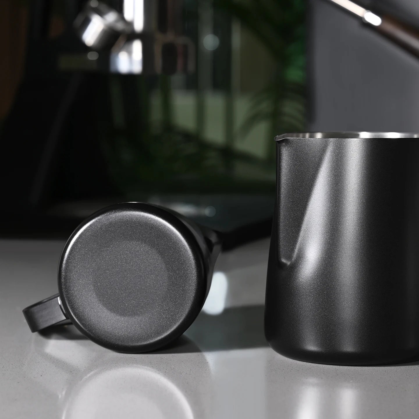A matte black stainless steel milk pitcher and matching handle are shown on a reflective light gray surface, with a blurred dark interior background.