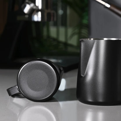 A matte black stainless steel milk pitcher and matching handle are shown on a reflective light gray surface, with a blurred dark interior background.