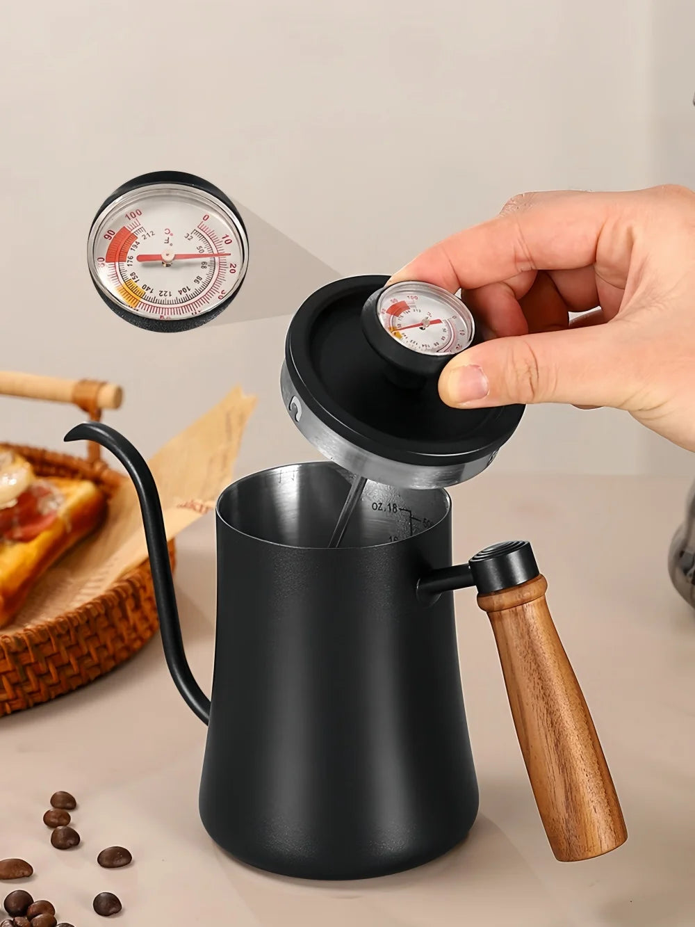 A matte black pour-over coffee maker with a wooden handle and a lid being lifted by a hand, showing a built-in dial thermometer. A close-up of the dial is shown in the upper left, with coffee beans and a slice of toast on a beige tabletop.
