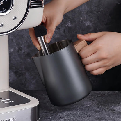 A matte black milk pitcher with a light wood handle being filled with steam from a white and stainless steel espresso machine, held by two hands against a dark textured background.