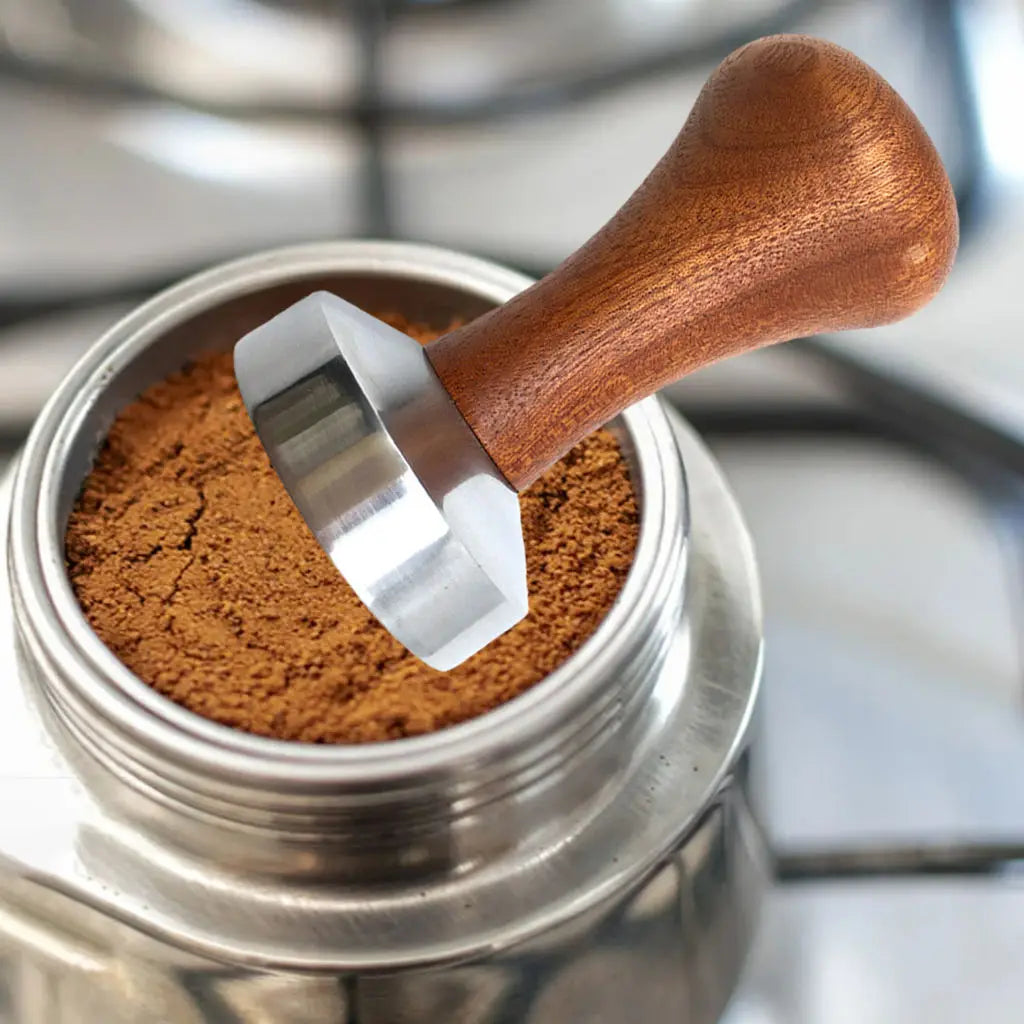 Wooden handle coffee tamp pressing coffee grounds into a portafilter basket.