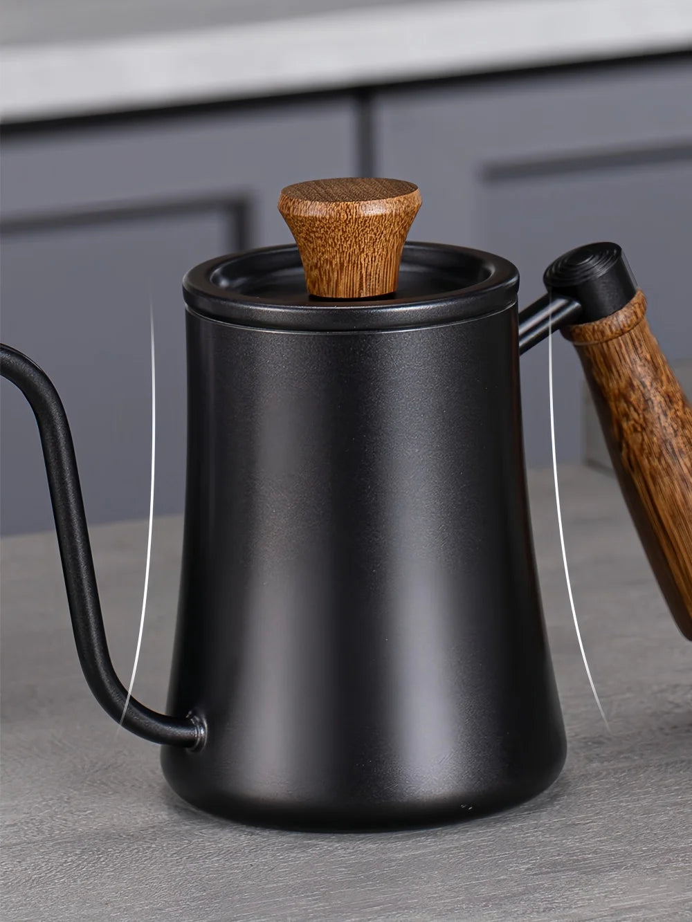 Matte black gooseneck kettle with a wooden handle and wooden lid knob, shown on a gray countertop with a blurred kitchen background.