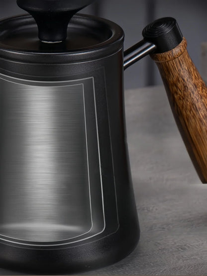 Close-up of a black metal kettle or teapot with a brushed silver front panel, black lid, and a brown wooden handle on a gray surface.