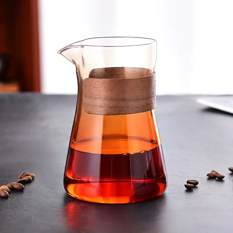 Glass carafe with wooden lid filled with red liquid on a dark surface with coffee beans.