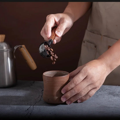 Person pouring coffee beans into a small brown ceramic cup with a metal scoop.
