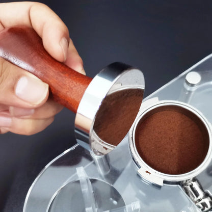 Hand using a wooden coffee tamper to compress coffee grounds in a filter basket.