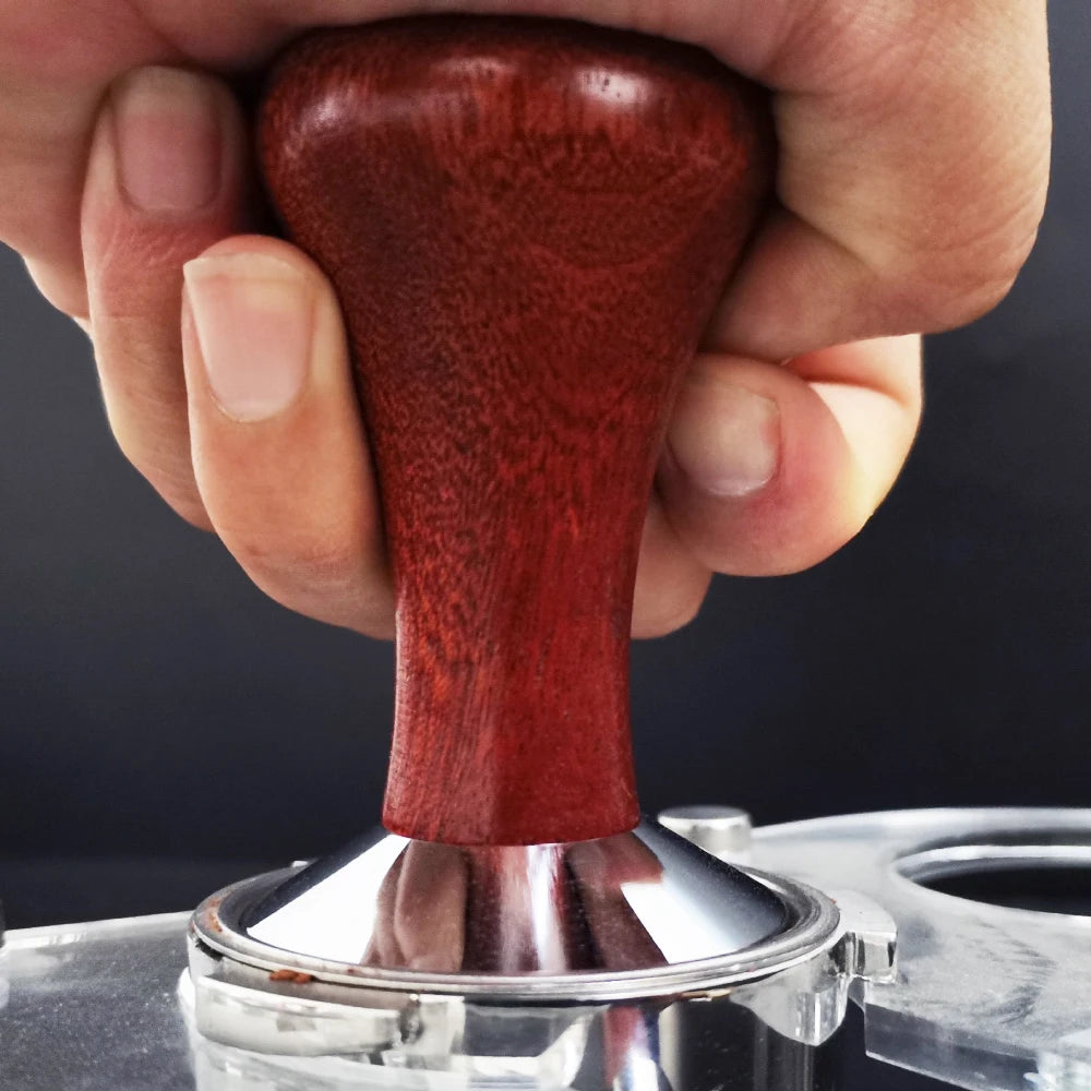 Hand using a red coffee tamp to compress grounds in a silver tamper against a dark background