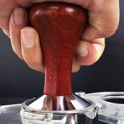 Hand using a red coffee tamp to compress grounds in a silver tamper against a dark background