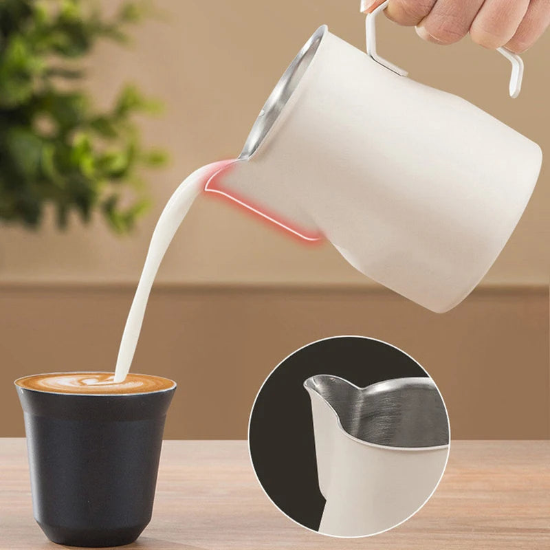 A hand holds a matte white milk pitcher with a stainless steel pouring spout, pouring steamed milk into a black cup of coffee. A circular inset shows a close-up of the pitcher's stainless steel pouring lip.