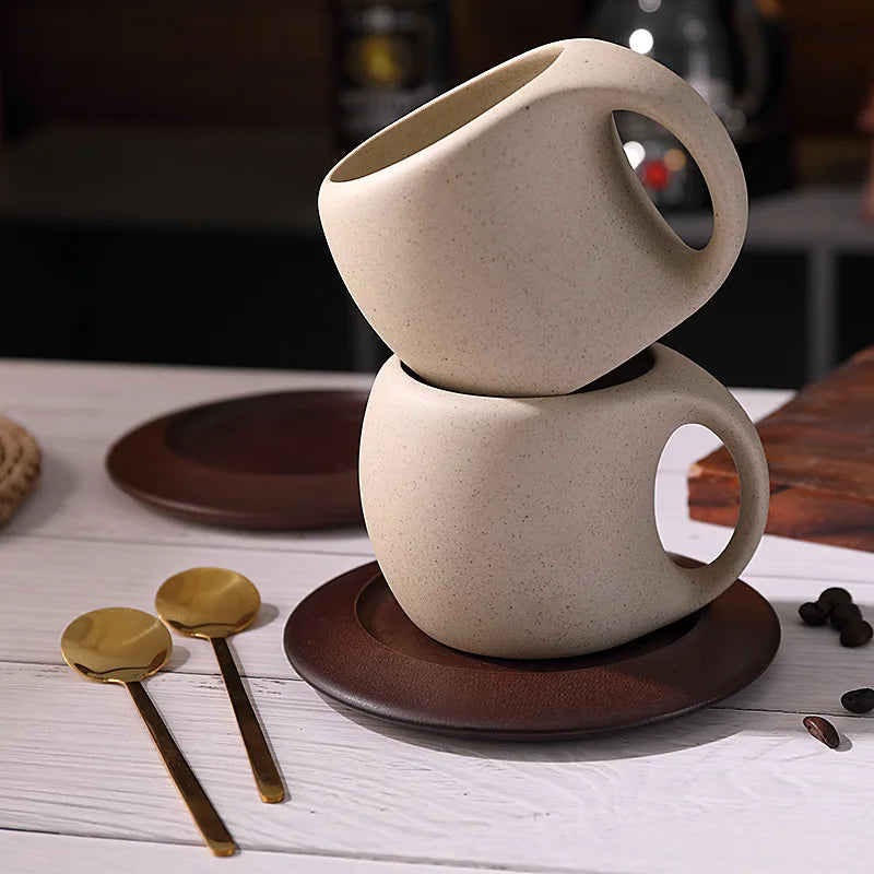 Two stacked ceramic coffee cups on a wooden coaster with gold spoons and coffee beans on a wooden table.