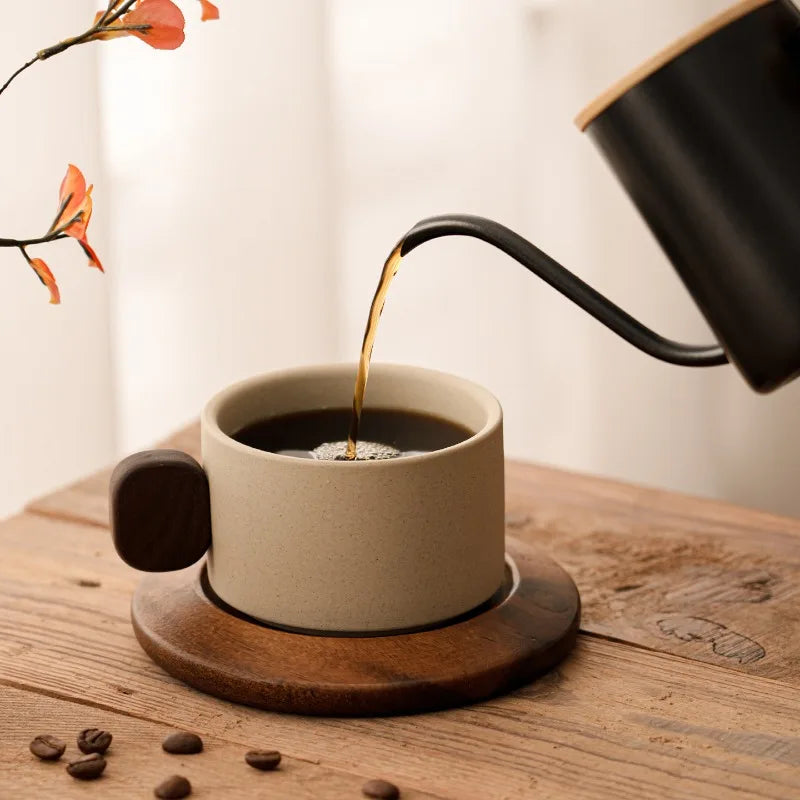 Black coffee being poured from a kettle into a beige mug on a wooden surface with scattered coffee beans.