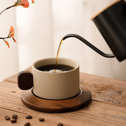 Black coffee being poured from a kettle into a beige mug on a wooden surface with scattered coffee beans.