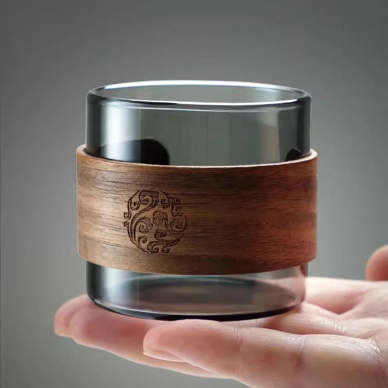 Clear glass candle holder with wooden sleeve held in a hand against a gray background