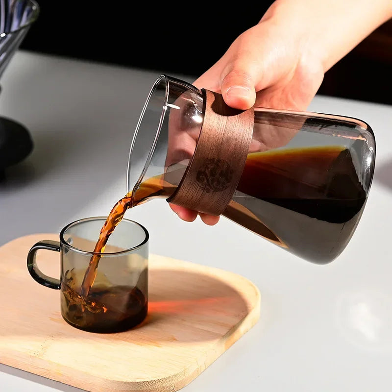 Person pouring coffee from a glass carafe with wooden handle into a glass mug on a light surface.