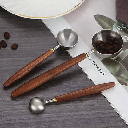 Three metal coffee scoop tools with brown wooden handles and gold-tone connectors, arranged on a gray wood surface with coffee beans and a white printed sheet in the background.
