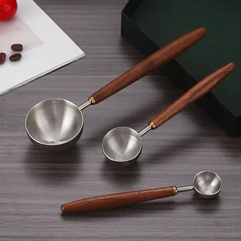 Three stainless steel tea ladles with brown wooden handles and gold-toned metal connectors, arranged on a dark wood tabletop with a notebook, coffee beans, and a box in the background.