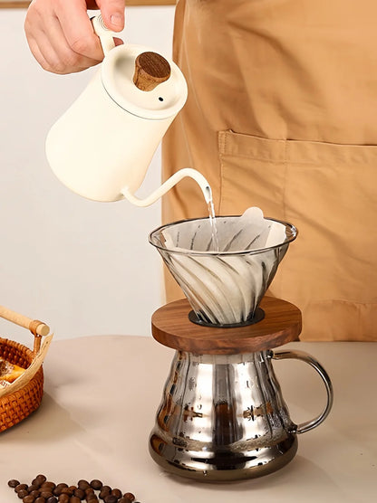 A person in a tan apron pours hot water from a white ceramic pour-over kettle into a glass cone filter sitting on a wooden ring, with a stainless steel carafe below and coffee beans on a tabletop.
