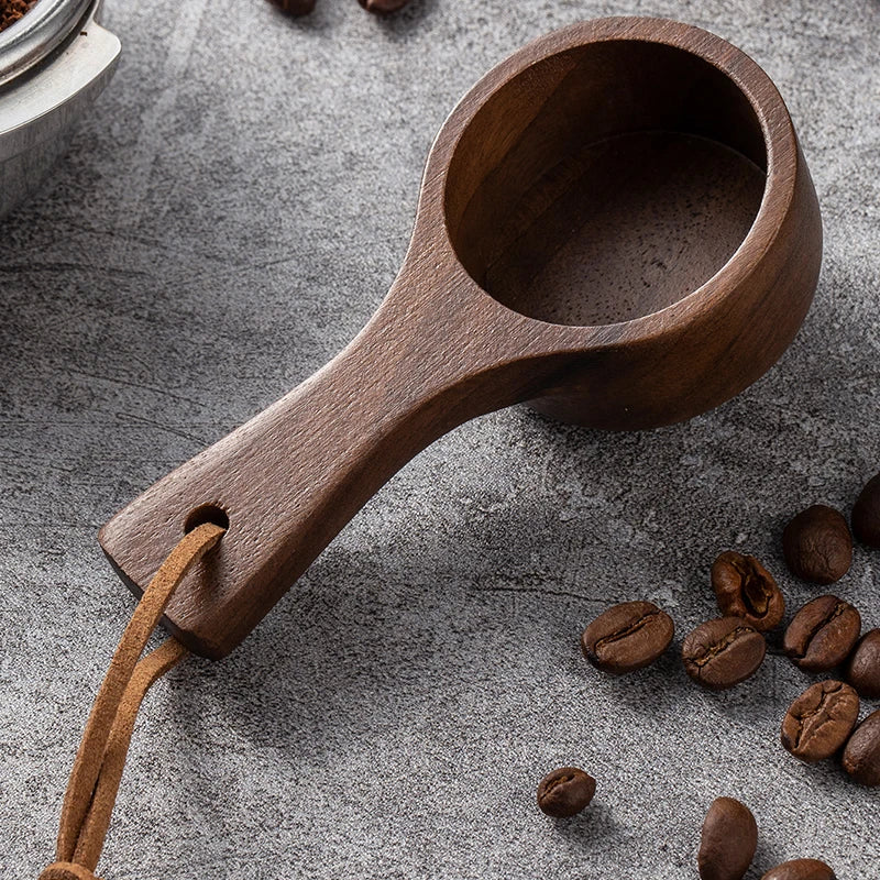 Wooden coffee scoop with a leather strap on a gray surface with scattered coffee beans.