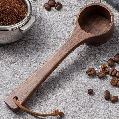 A dark brown wooden coffee scoop with a hanging loop, shown on a textured gray surface with coffee beans and a metal coffee bean hopper.